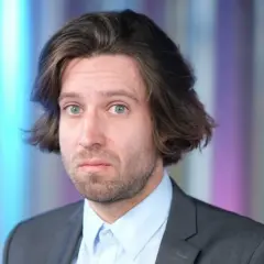 Michał Kempa stand up, w garniturze i jasnej koszuli, z burzą brązowych włosów i niebieskimi oczami, na tle kolorowych pasów.