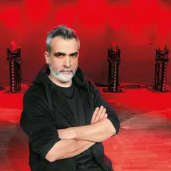 Abelard Giza stand up, pewny siebie, z rękami skrzyżowanymi na piersi, na tle czerwonych świateł scenicznych.