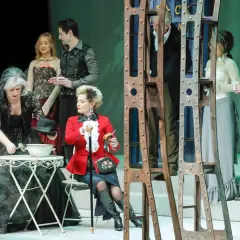 Scena z przedstawienia "Teatr kwadrat za jakie grzechy". Kobieta w czarnej sukni nalewa coś z misy, obok siedzi dama w czerwonym żakiecie.