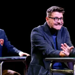 Dwóch aktorów na scenie podczas spektaklu "Stowarzyszenie Umarłych Poetów". Młody mężczyzna w garniturze i starszy z okularami, obaj siedzą przy stołach.