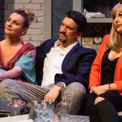 Trzy osoby siedzą na kanapie. Kobieta w kolorowej sukience, mężczyzna w marynarce i kobieta w pomarańczowej marynarce. Spektakl "Wiesz, że wiem".