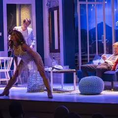 Scena teatralna: kobieta w białej, ażurowej sukience i mężczyzna w marynarce siedzą na kanapie.