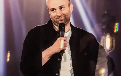 Marcin Zbigniew Wojciech w swoim stand-upie, trzyma mikrofon, a światła sceniczne tworzą tęczowe refleksy.