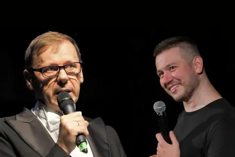 Dwóch mężczyzn z mikrofonami na scenie. Jeden w garniturze, drugi w czarnej koszulce. Widać, że socha stand up.