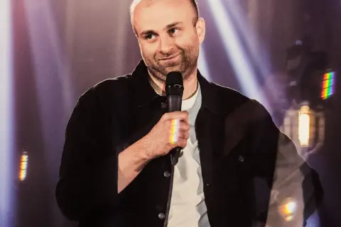 Marcin Zbigniew Wojciech w swoim stand-upie, trzyma mikrofon, a światła sceniczne tworzą tęczowe refleksy.