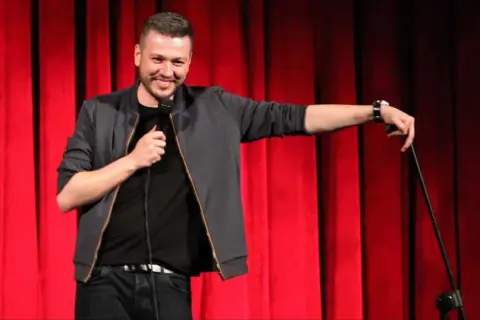 Mateusz Socha na scenie z mikrofonem, na tle czerwonej kurtyny.
