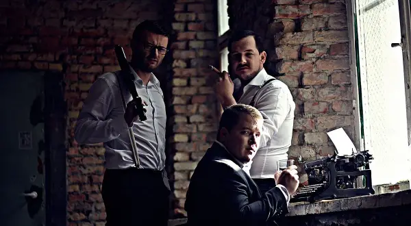 Trzech mężczyzn w białych koszulach i szelkach, jeden z cygarem, obok maszyny do pisania.