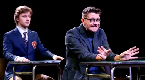 Dwóch aktorów na scenie podczas spektaklu "Stowarzyszenie Umarłych Poetów". Młody mężczyzna w garniturze i starszy z okularami, obaj siedzą przy stołach.