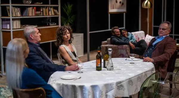 Scena z przedstawienia "Kto chce być żydem", teatr współczesny. Przy stole siedzą ludzie, w tle para na kanapie.