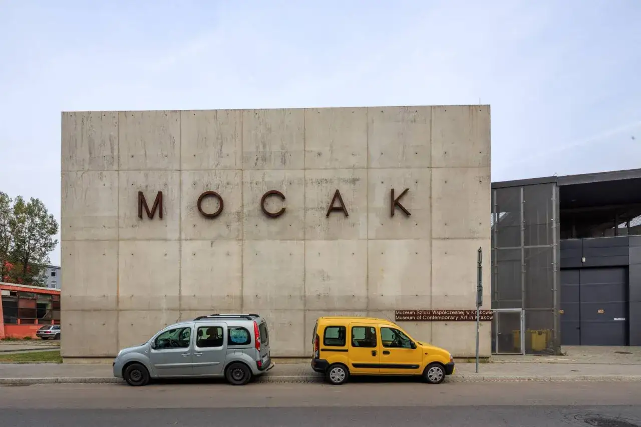 Betonowa fasada muzeum sztuki wsp&oacute;łczesnej w Krakowie MOCAK z literami nazwy. Dwa samochody stoją przed budynkiem.