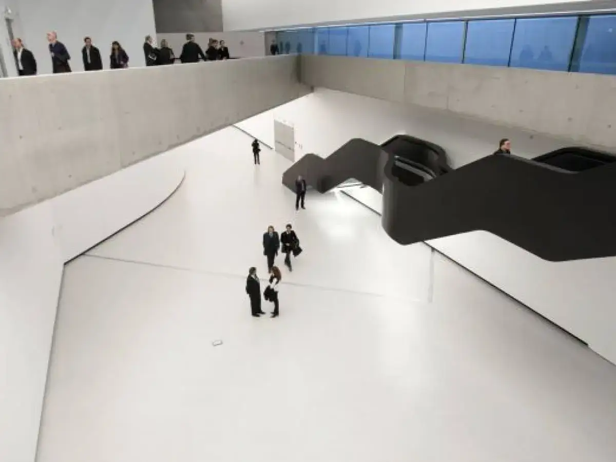 Wnętrze rzymskiego muzeum sztuki nowoczesnej z ludźmi spacerującymi po białej przestrzeni i czarnymi, falistymi schodami.