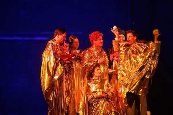 Aktorzy w złotych szatach, trzymający filiżanki i książki, tworzą scenę z najlepszych spektakli teatralnych.