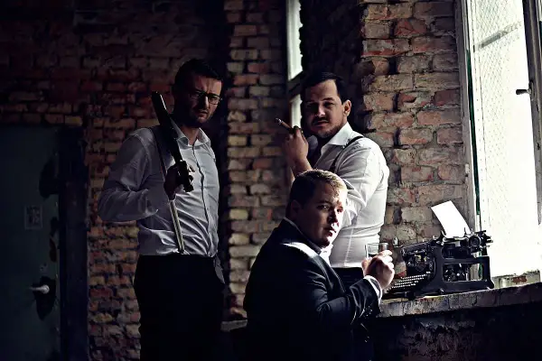 Trzech mężczyzn w białych koszulach i szelkach, jeden z cygarem, obok maszyny do pisania.
