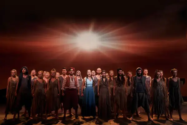 Chór w "Aidzie" w Teatrze Roma, spektakl trwa. Grupa ludzi w strojach z epoki na tle dramatycznego, rozświetlonego nieba.