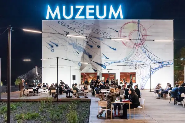 Wieczorem muzeum sztuki nowoczesnej nad Wisłą tętni życiem. Ludzie siedzą przy stolikach na zewnątrz, podziwiając abstrakcyjną grafikę na ścianie budynku.