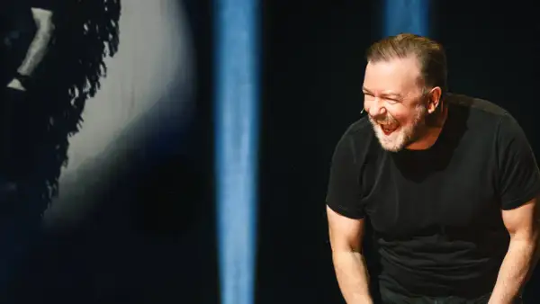 Ricky Gervais w swoim stand upie śmieje się do rozpuku, w czarnym t-shircie, na tle niebieskich świateł.