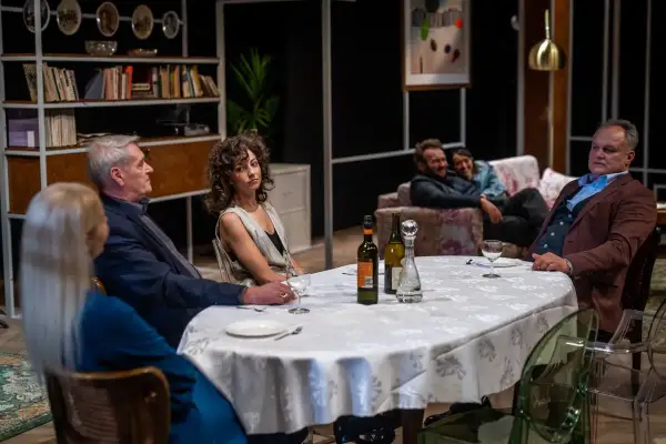 Scena z przedstawienia "Kto chce być żydem", teatr współczesny. Przy stole siedzą ludzie, w tle para na kanapie.